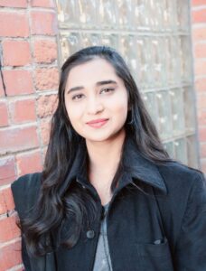 A woman with softly curled dark hair stands in front of a brick wall. She wears a dark colored jacket and has a closed-lip smile on her face.