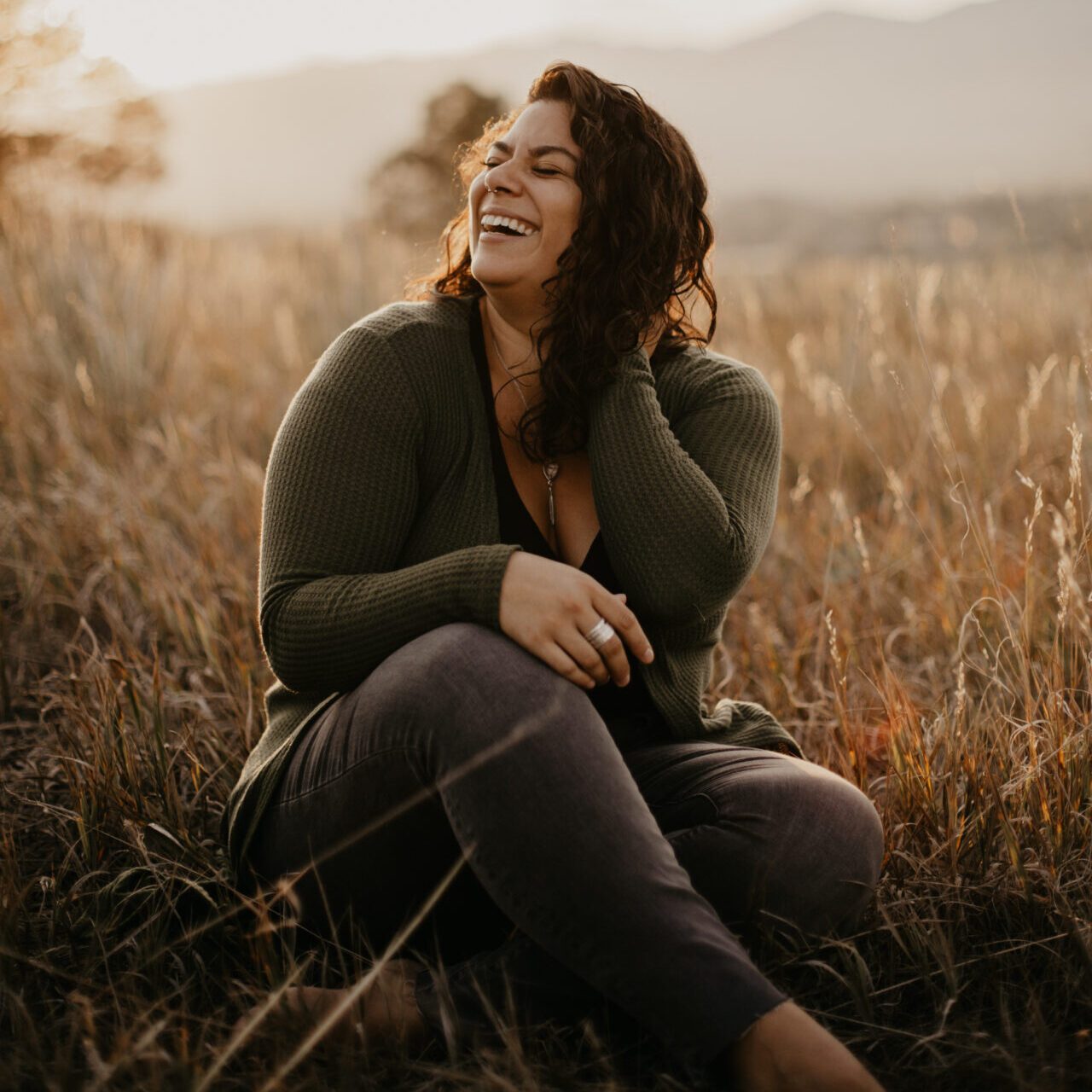 A woman in a green top with brown hair laughs in a field with the sun behind her.